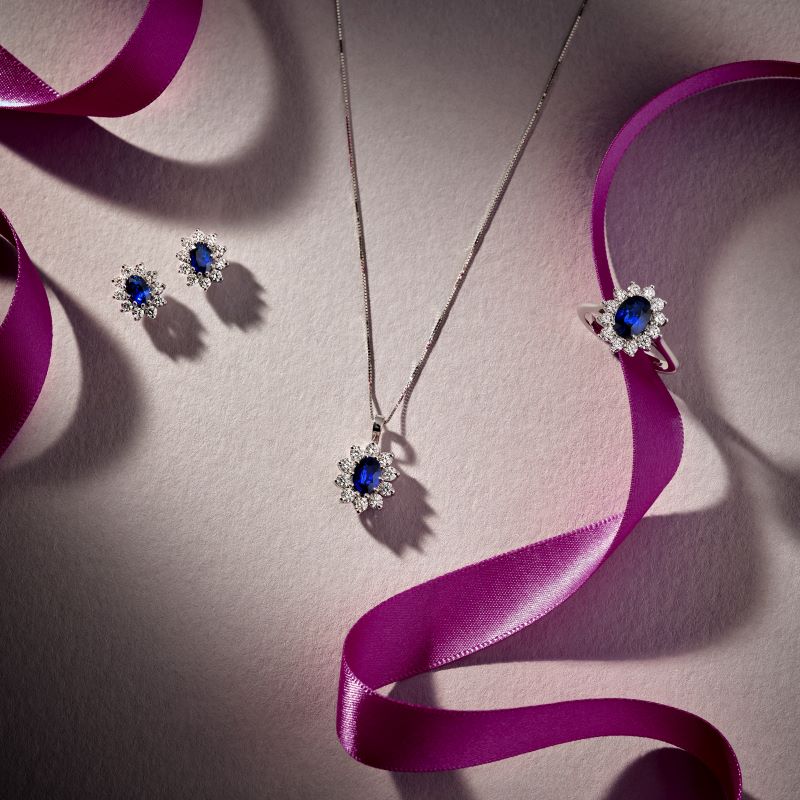 18ct white gold oval blue sapphire and diamond earrings, necklace and ring, each surrounded by round brilliant diamonds in a claw setting, displayed with purple ribbon.