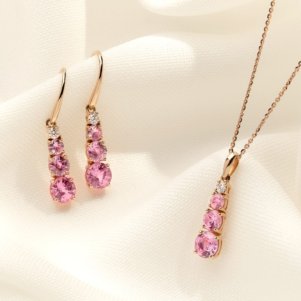 Pink sapphire and diamond drop earrings with a matching pendant in 18ct rose gold, each featuring three graduated round pink sapphires and a brilliant cut diamond, displayed on cream fabric.