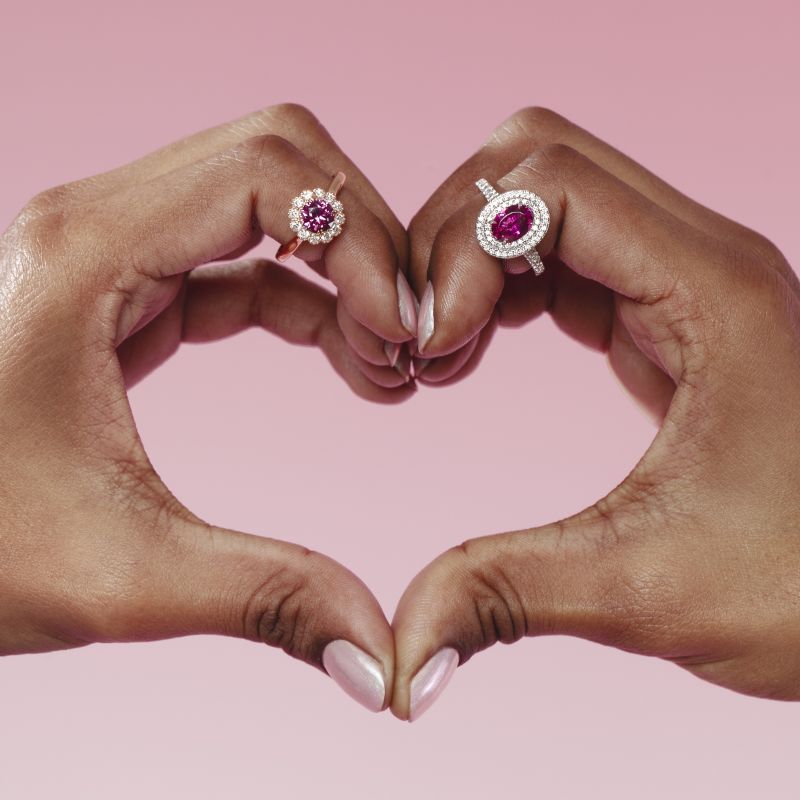 Pink tourmaline and rhodolite garnet rings worn on hands forming a heart, featuring an oval pink tourmaline with a double diamond halo in platinum and a round rhodolite garnet with a diamond halo in 1